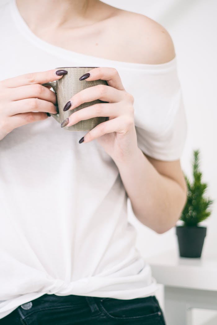 services-02 Close-up of a woman with manicured nails holding a mug in a bright, minimalist setting.